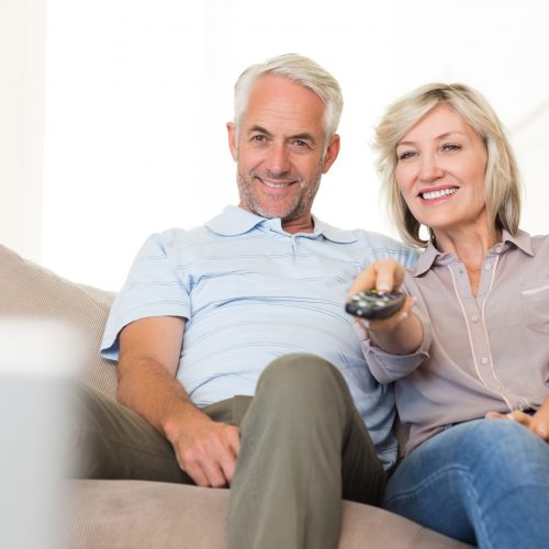 Happy mature couple watching tv on sofa at home